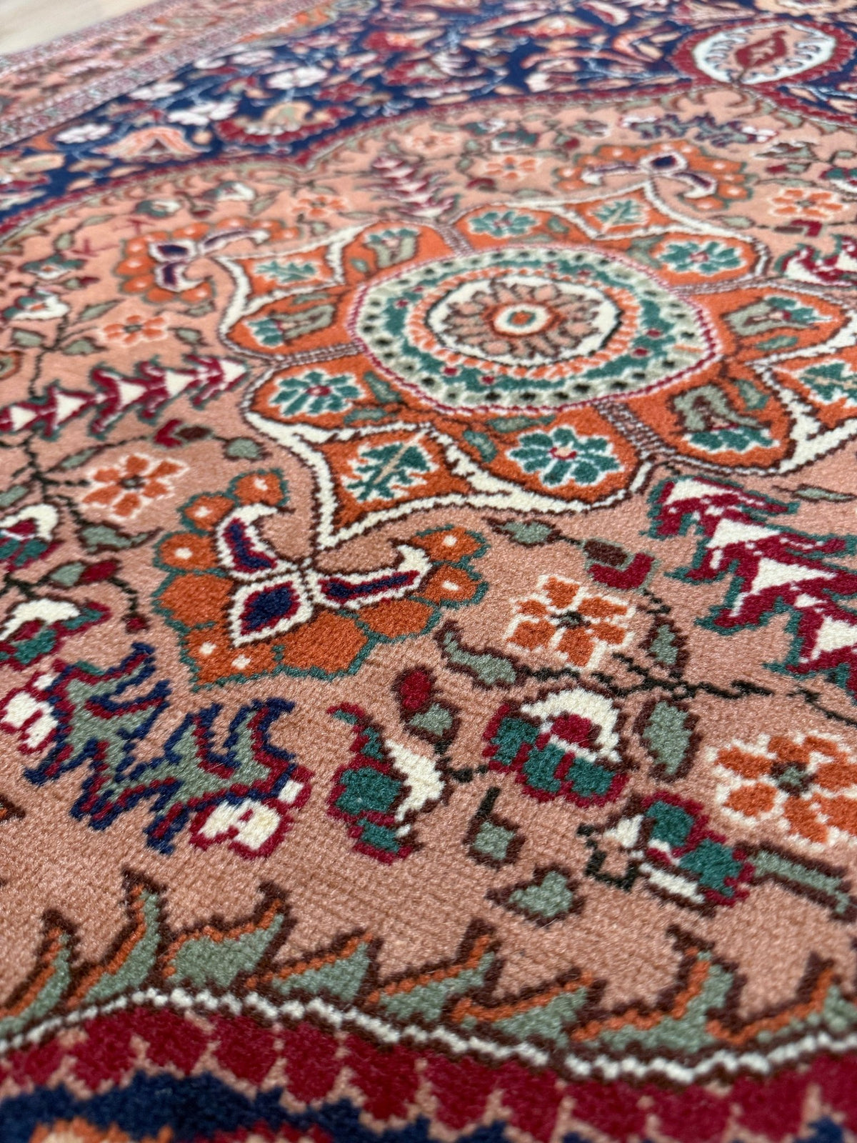 Close-up of an ornate traditional rug with a dusty pink field, featuring intricate floral and medallion patterns in orange, teal, red, and cream. 5x7 Navy Kayseri Turkish rug with peach medallion, floral vines, handmade wool from Indigo Rugs Burlingame Bay Area.