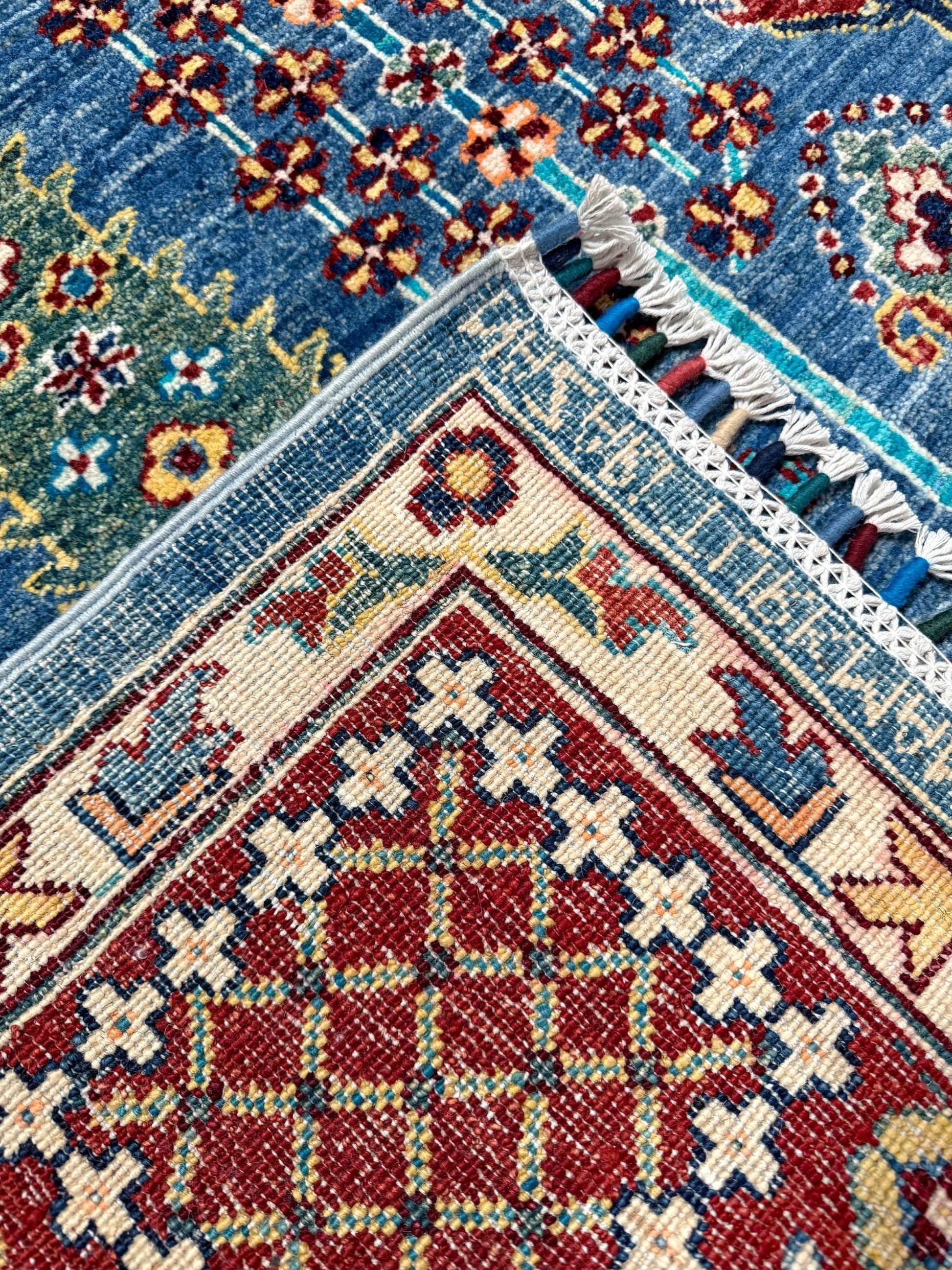 Close-up of a patterned area rug corner showing a red central field with cream geometric motifs, bordered by blue and green floral sections with white tassels.