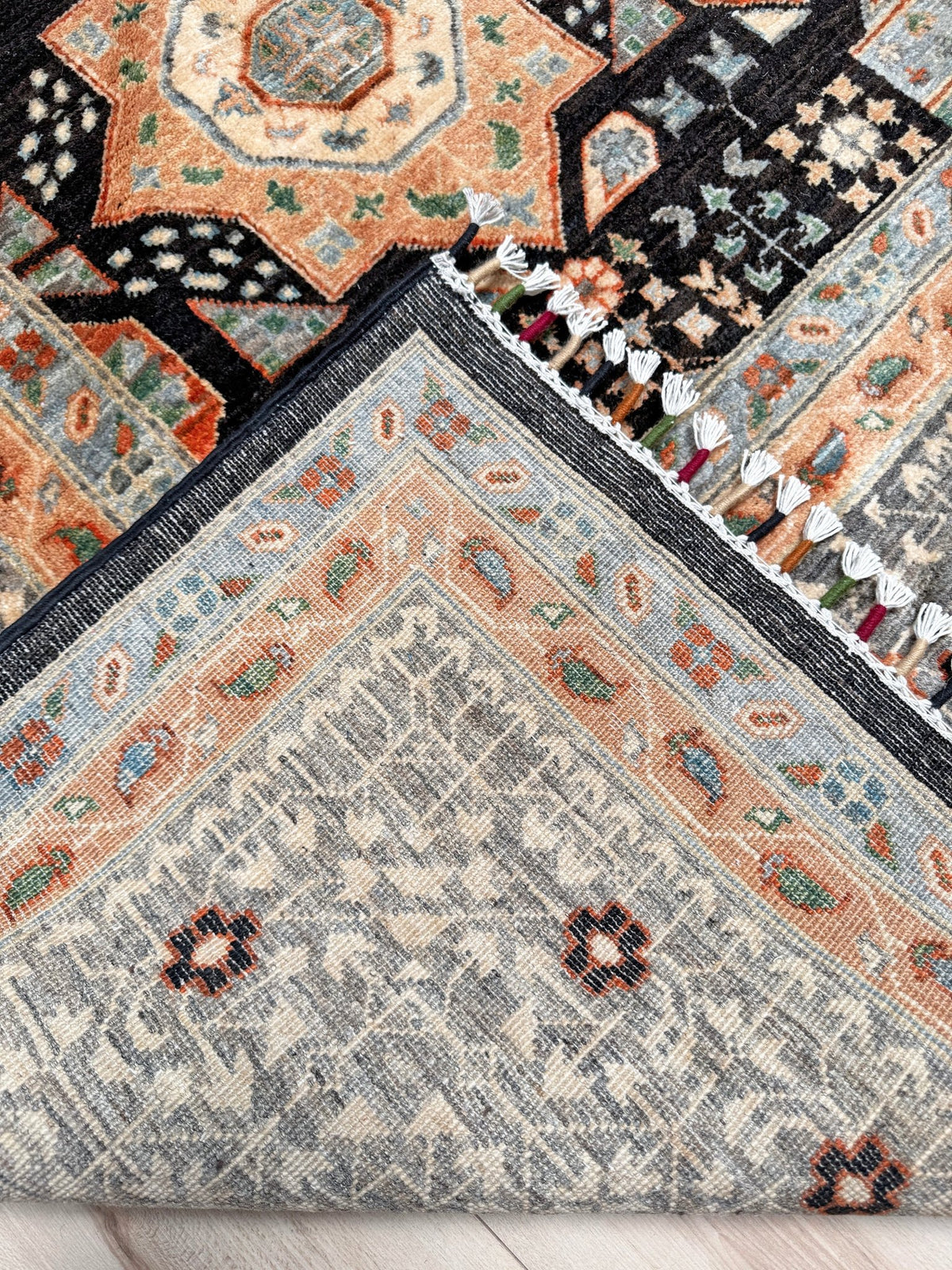 A close-up of a patterned area rug corner showing a gray central field, peach and blue borders, black sections, and white fringe tassels on a light wood floor.