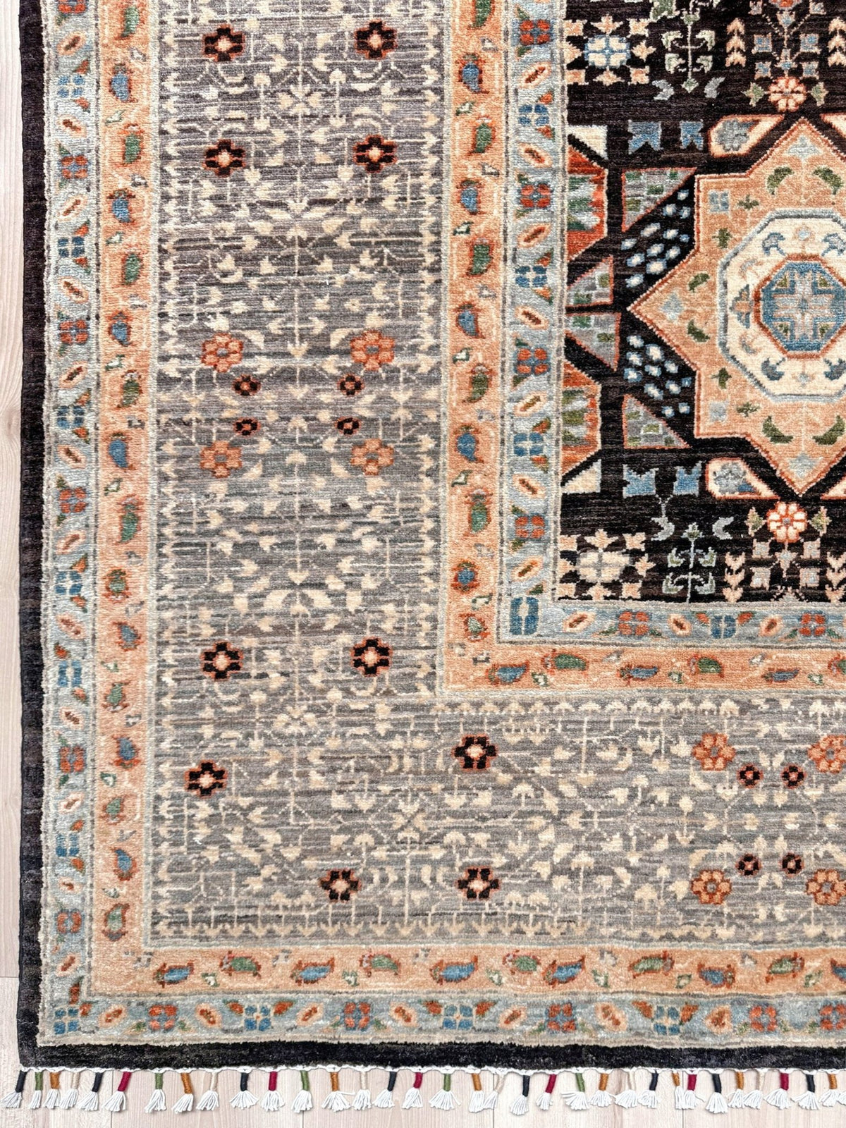 A close-up of a patterned area rug showing the corner and border details. The rug features a gray field with small floral motifs, a peach and beige inner border, and a dark navy outer border with cream fringe tassels. 10x14 Charcoal and peach Sultanate Mamluk rug with large central medallion, geometric floral motifs, handmade Turkish wool from Indigo Rugs Burlingame, SF Bay Area.