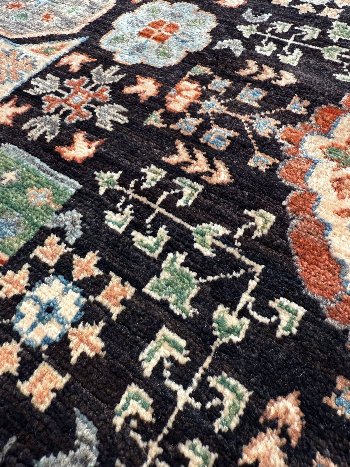 A close-up of a traditional patterned rug with a black base and intricate floral motifs in green, orange, beige, and blue. 10x14 Charcoal and peach Sultanate Mamluk rug with large central medallion, geometric floral motifs, handmade Turkish wool from Indigo Rugs Burlingame, SF Bay Area.