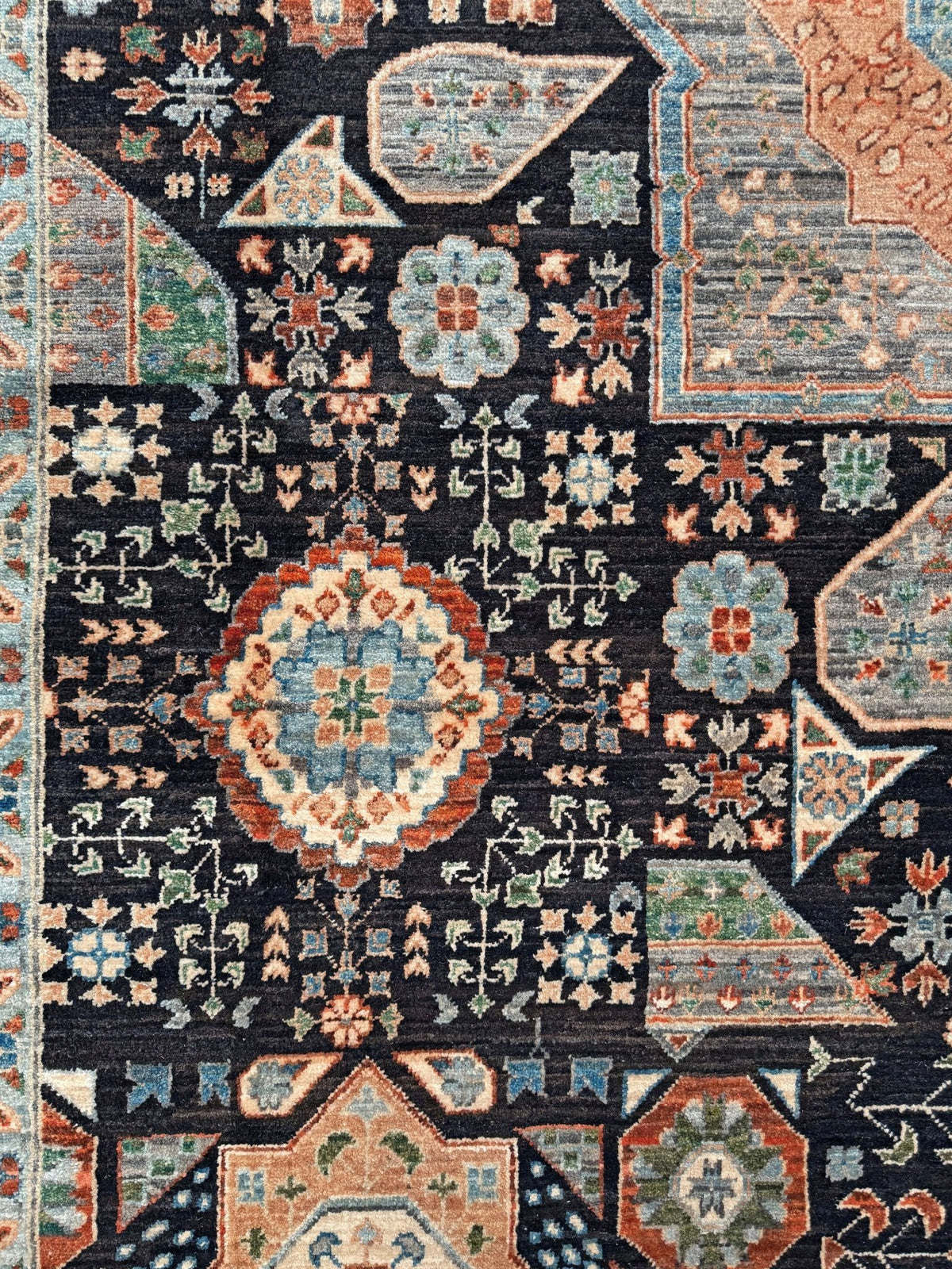 A close-up of a traditional patterned rug featuring a dark charcoal background with intricate medallions and floral motifs in muted blue, terracotta, beige, and sage green. 10x14 Charcoal and peach Sultanate Mamluk rug with large central medallion, geometric floral motifs, handmade Turkish wool from Indigo Rugs Burlingame, SF Bay Area.