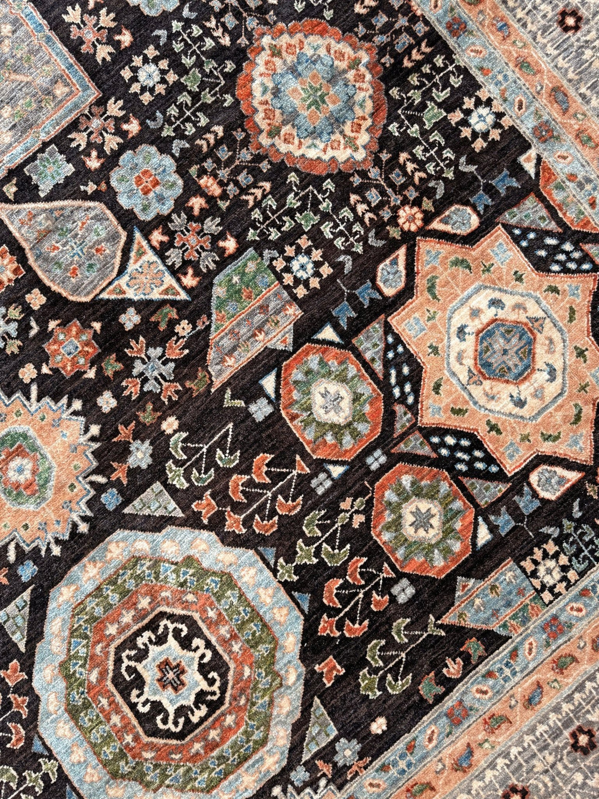 A close-up of a patterned area rug featuring a dark charcoal black base with intricate geometric and floral motifs in terracotta, beige, light blue, and muted green. 10x14 Charcoal and peach Sultanate Mamluk rug with large central medallion, geometric floral motifs, handmade Turkish wool from Indigo Rugs Burlingame, SF Bay Area.