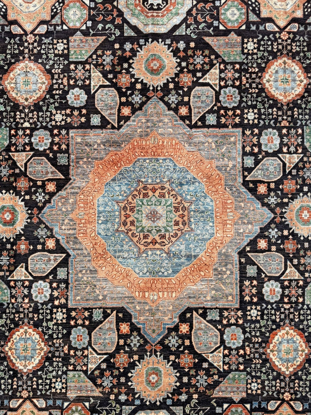A close-up of an ornate area rug featuring a central medallion and intricate geometric floral motifs in muted blue, terracotta, beige, and cream on a dark charcoal background. 10x14 Charcoal and peach Sultanate Mamluk rug with large central medallion, geometric floral motifs, handmade Turkish wool from Indigo Rugs Burlingame, SF Bay Area.