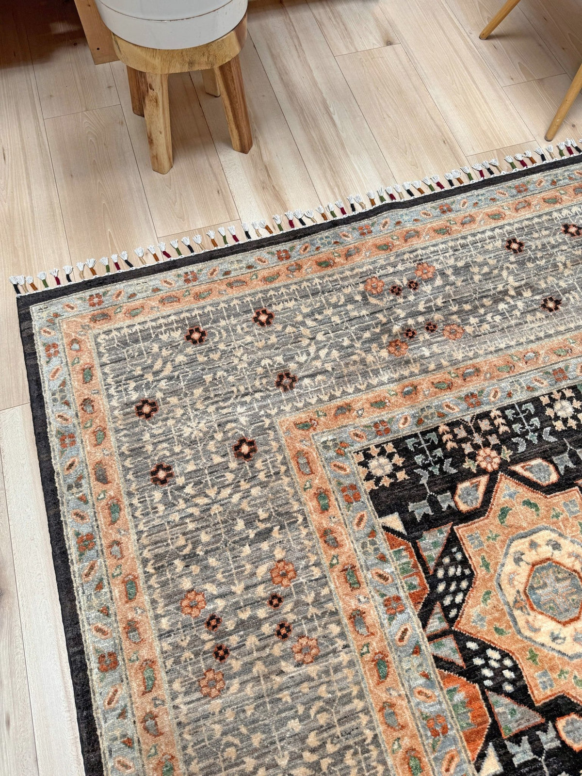 A close-up of a traditional area rug with a distressed gray field, peach and orange floral motifs, and a black border, shown on a light wood floor. 10x14 Charcoal and peach Sultanate Mamluk rug with large central medallion, geometric floral motifs, handmade Turkish wool from Indigo Rugs Burlingame, SF Bay Area.
