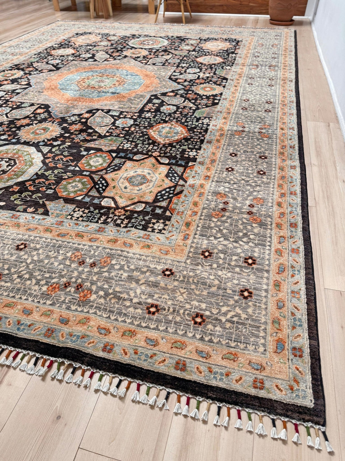 A traditional oriental-style area rug with a complex geometric and floral pattern, shown laid out on a light wood floor. The rug features a dark charcoal/black base with intricate borders and medallions in beige, orange, light blue, and gray, finished with a white and multi-colored fringe.