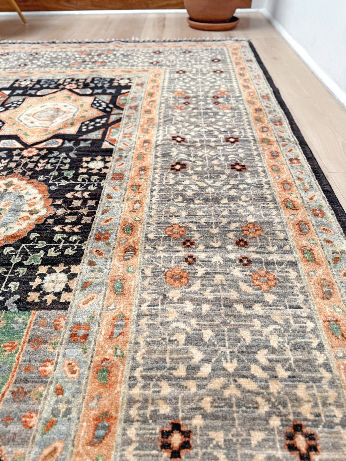 A traditional oriental-style area rug with a distressed gray field, orange and beige borders, and a black central panel, shown on a light wood floor. 10x14 Charcoal and peach Sultanate Mamluk rug with large central medallion, geometric floral motifs, handmade Turkish wool from Indigo Rugs Burlingame, SF Bay Area.