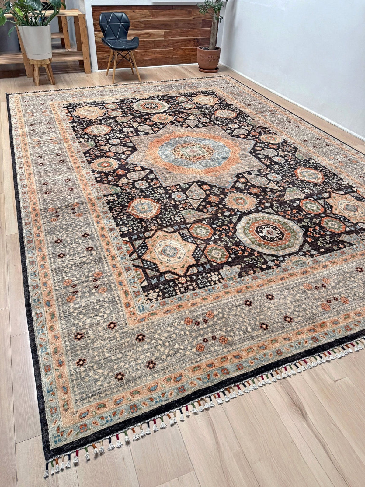 A large rectangular area rug with an intricate traditional medallion pattern, shown on a light wood floor in a modern room setting. 10x14 Charcoal and peach Sultanate Mamluk rug with large central medallion, geometric floral motifs, handmade Turkish wool from Indigo Rugs Burlingame, SF Bay Area.