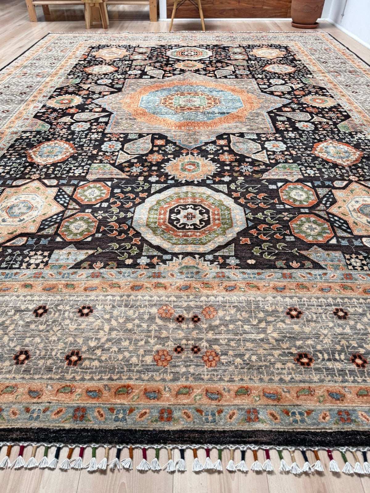 A large rectangular area rug with an intricate traditional medallion pattern, shown laid out on a light wood floor in a room. The rug features a black central field, a beige and gray patterned border, and accents of orange, blue, and green in the medallions and floral motifs.