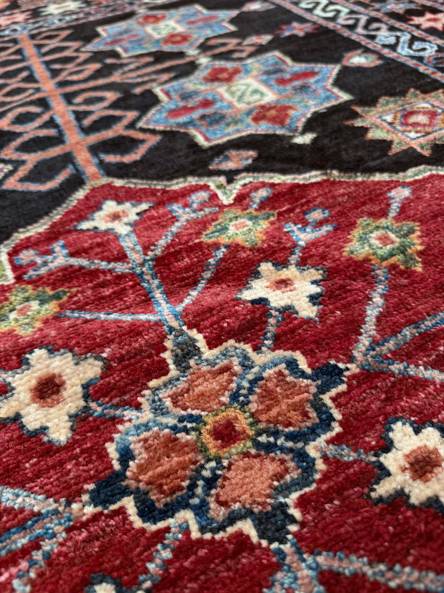 A close-up of a traditional patterned rug with a deep red field, cream floral motifs, and blue accents, transitioning into a black section with geometric designs.