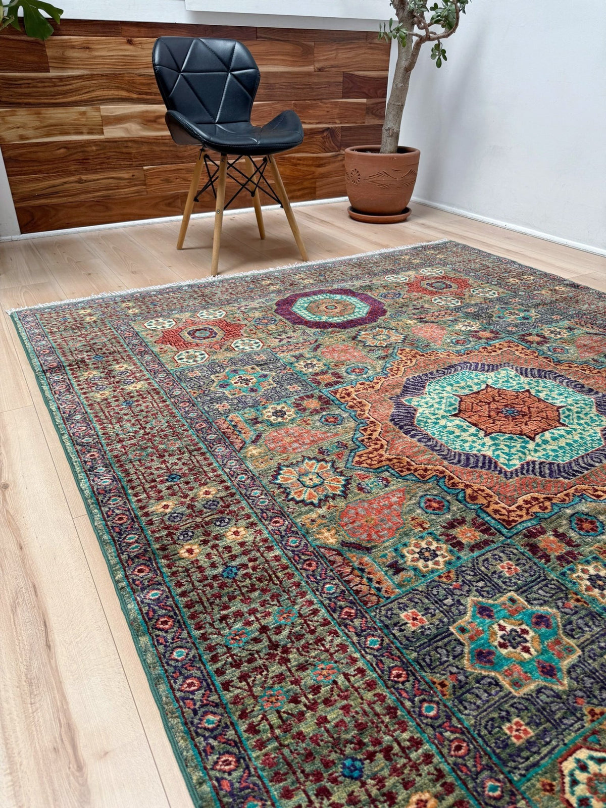 A large traditional area rug with an intricate multicolor pattern in shades of teal, rust, navy, and beige, styled on a light wood floor in a modern room setting with a black chair and a potted plant.