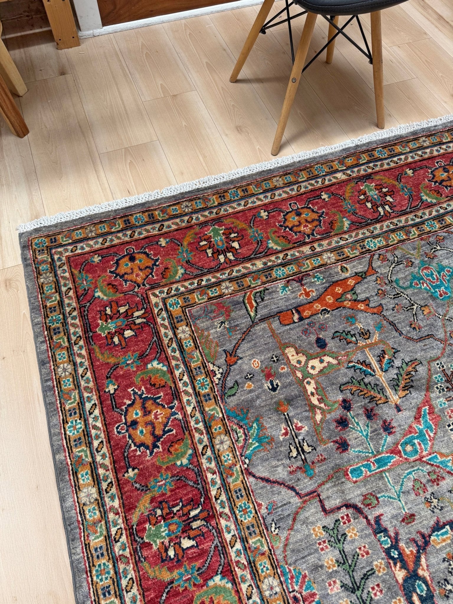 A traditional oriental area rug with a gray field and red border, shown on a light wood floor in a room setting with a chair partially visible.