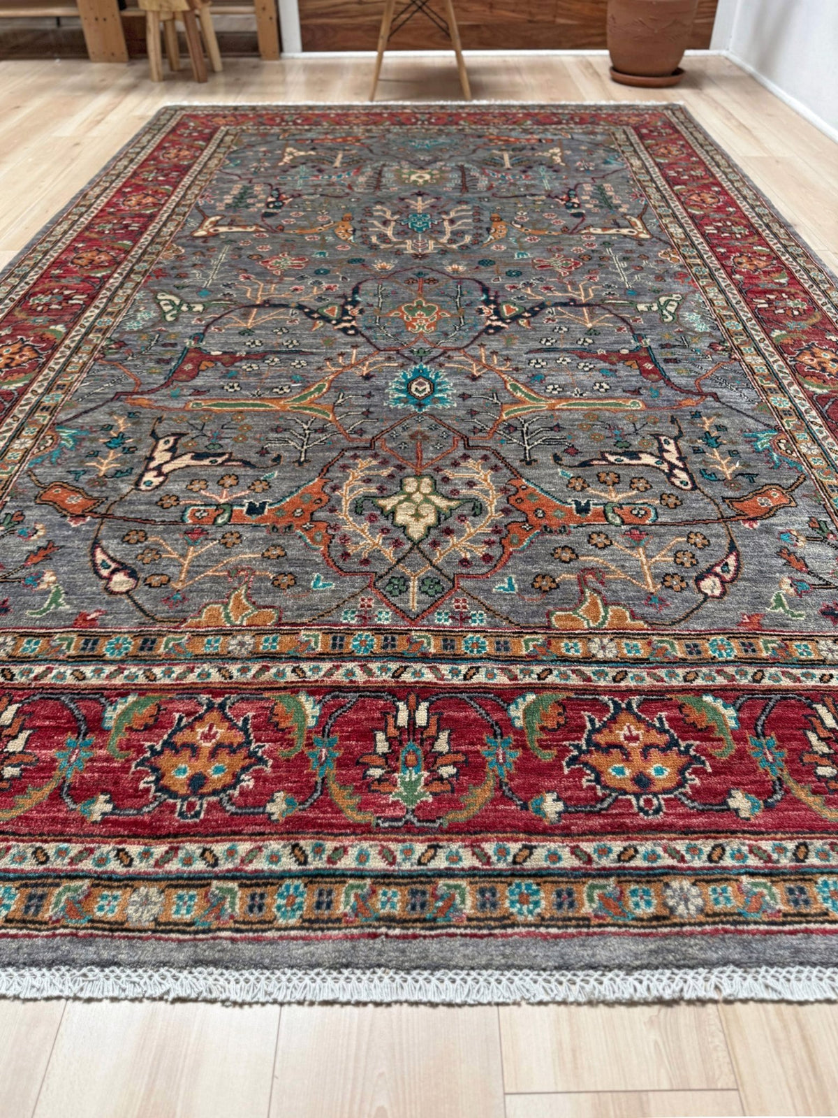 A traditional Persian-style area rug with a complex floral and geometric pattern, featuring a gray-blue field, red borders, and accents of orange, green, and beige, laid out on a light wood floor.