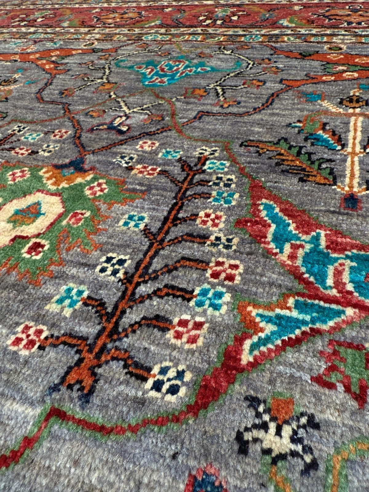 A close-up of a traditional handwoven rug with a gray base and intricate floral and vine patterns in red, blue, green, black, and cream.