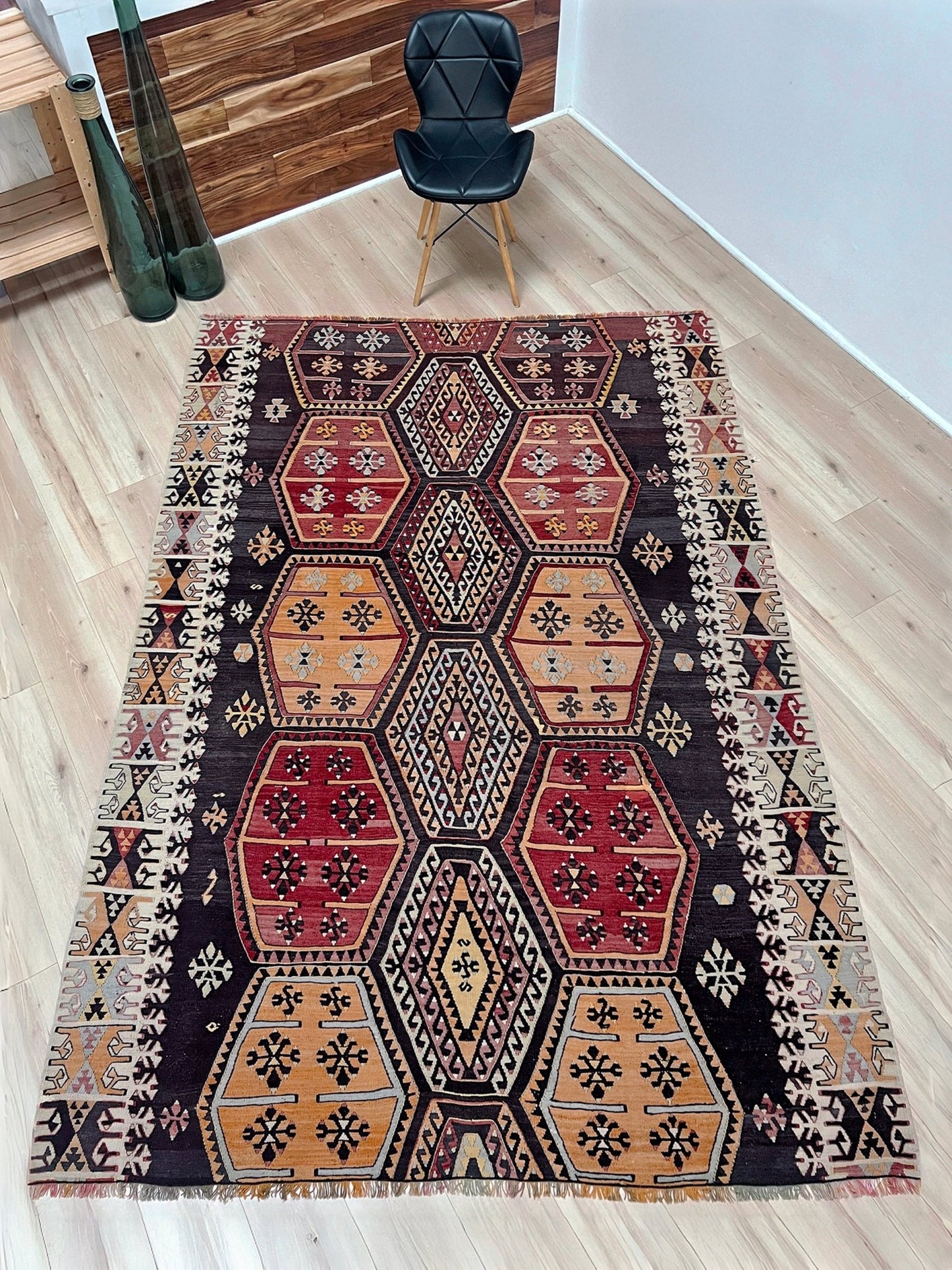 Patterned rug on a wooden floor with a chair and decor items in the background. Vintage Sarkisla Turkish kilim rug 6x9 with deep brown field and geometric medallions in madder red, camel, and ivory tones — handmade Anatolian flatweave carpet