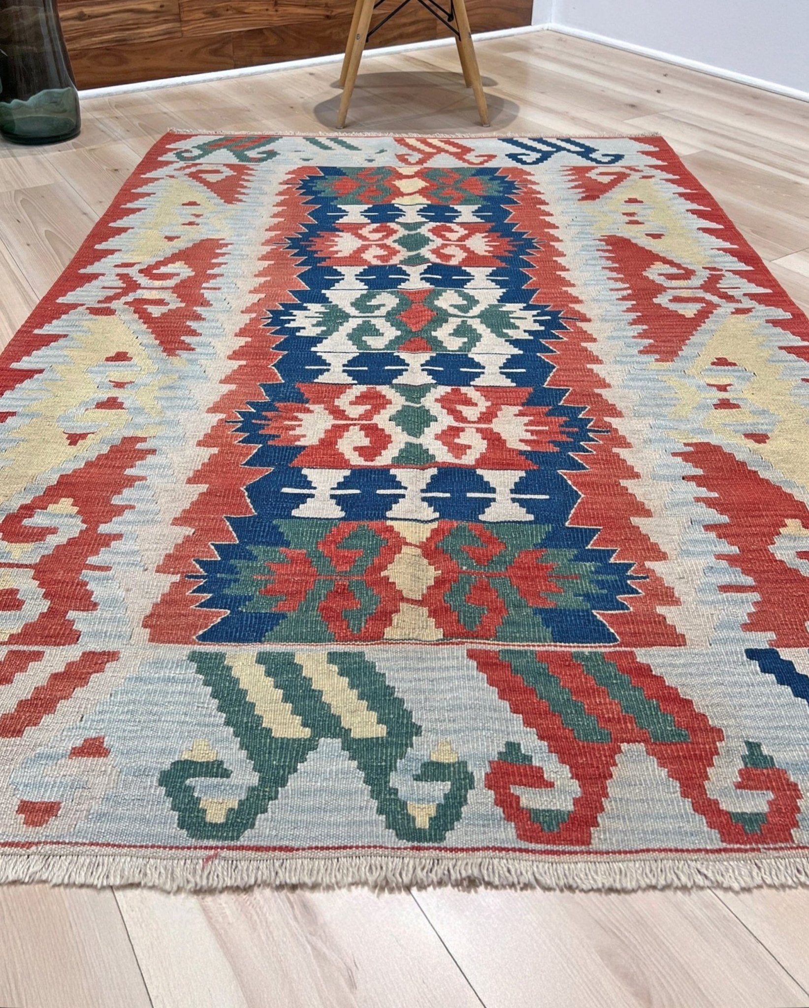 Colorful patterned rug on a wooden floor with a chair in the background. Woven turkish kilim rug with geometric pattern featuring red, blue, and beige colors. 