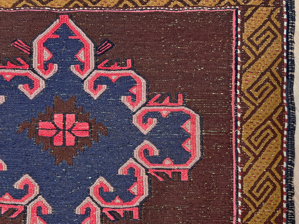 Close-up of a woven rug showing a dark brown field with a large blue medallion outlined in pink, plus a gold and brown geometric border on the right.