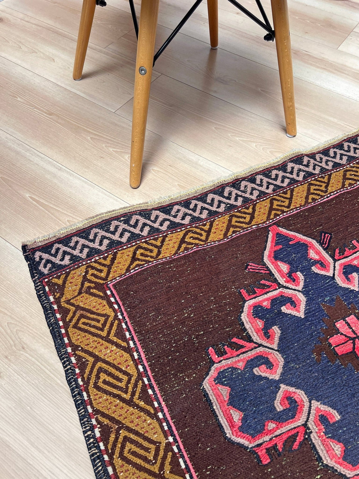 A close-up of a patterned area rug corner on a light wood floor, with the legs of a wooden chair visible at the top. The rug has a dark brown field, a blue central motif, and geometric borders in tan, cream, and pink.