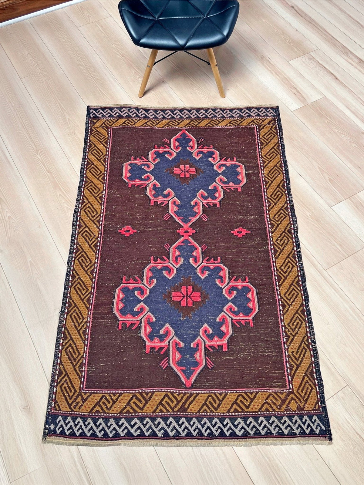 A rectangular vintage-style area rug with a dark brown field, featuring a central blue and pink geometric medallion motif, surrounded by a gold and navy geometric border. The rug is displayed on a light wood floor with a modern black leather chair partially visible at the top.