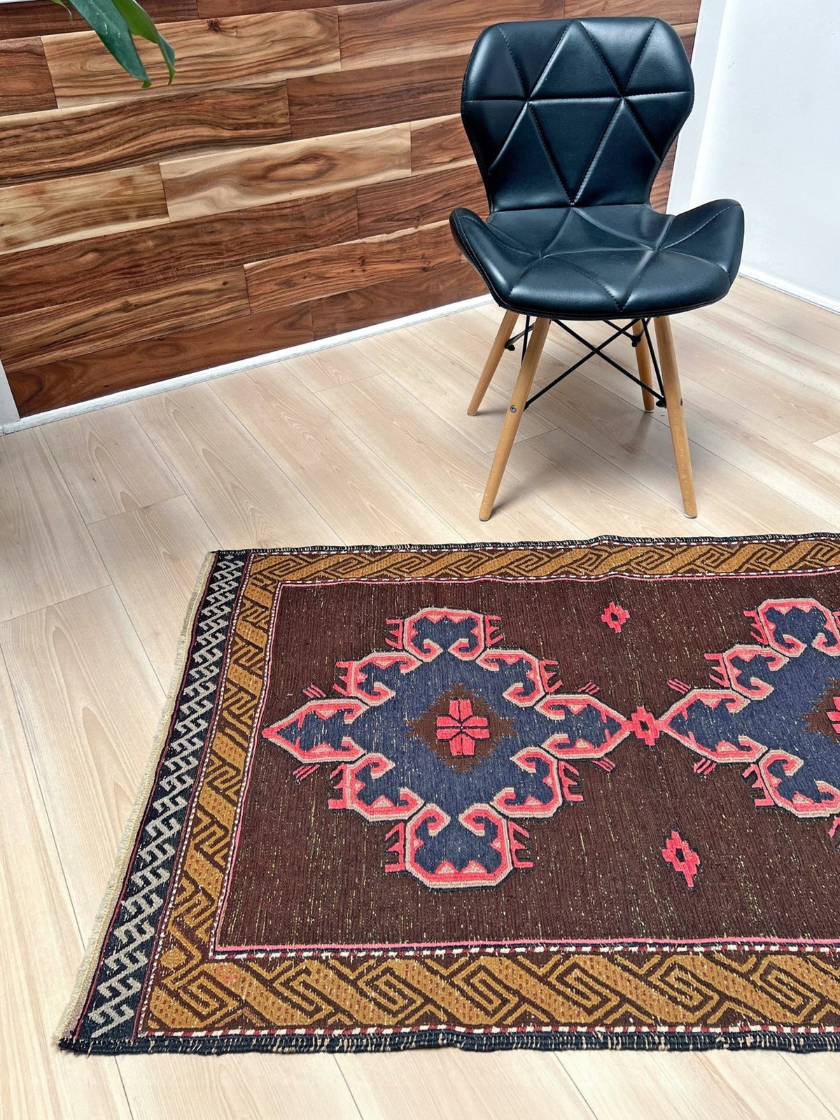 A rectangular area rug with a dark brown field, pink and blue geometric medallion pattern, and a tan Greek key border, shown on a light wood floor next to a black modern chair.