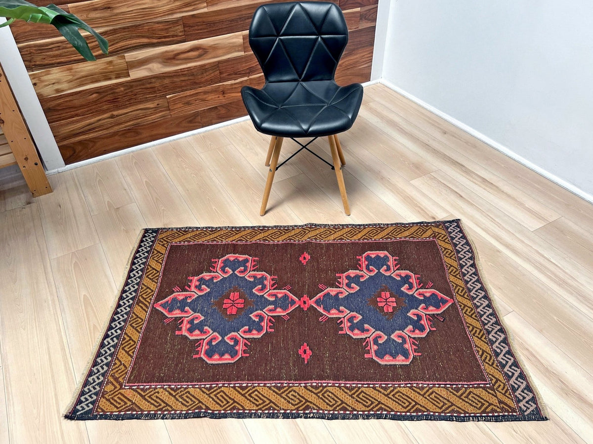 A rectangular vintage-style kilim rug with a brown background, blue medallions, and pink accents, styled on a light wood floor in a modern room with a black chair.