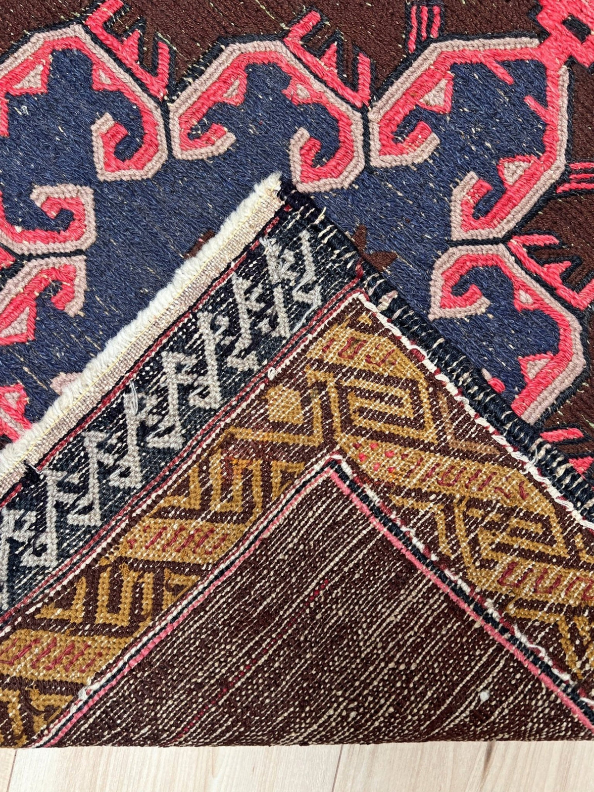A close-up of a patterned area rug showing multiple geometric and tribal-inspired sections in dark brown, navy blue, pink, beige, and gold, with visible woven texture and a light wood floor at the bottom.