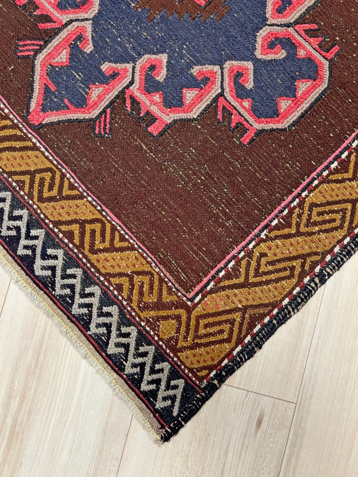 A close-up of a patterned area rug corner on a light wood floor, showing a dark brown field with blue and pink motifs, tan and black geometric borders, and fringe.