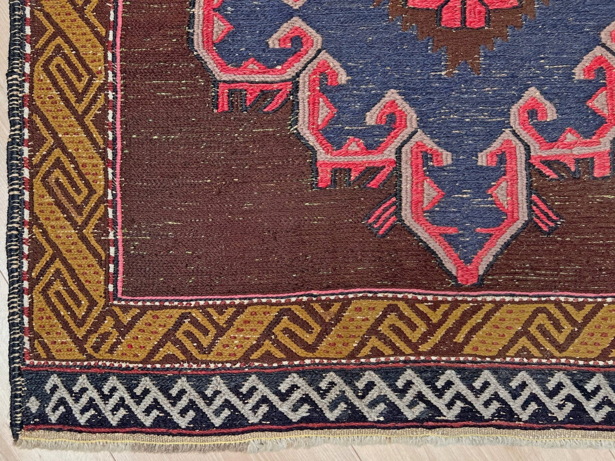 Close-up of a woven rug corner showing a dark brown field, a blue and pink central medallion motif, a mustard yellow geometric border, and a navy patterned fringe.