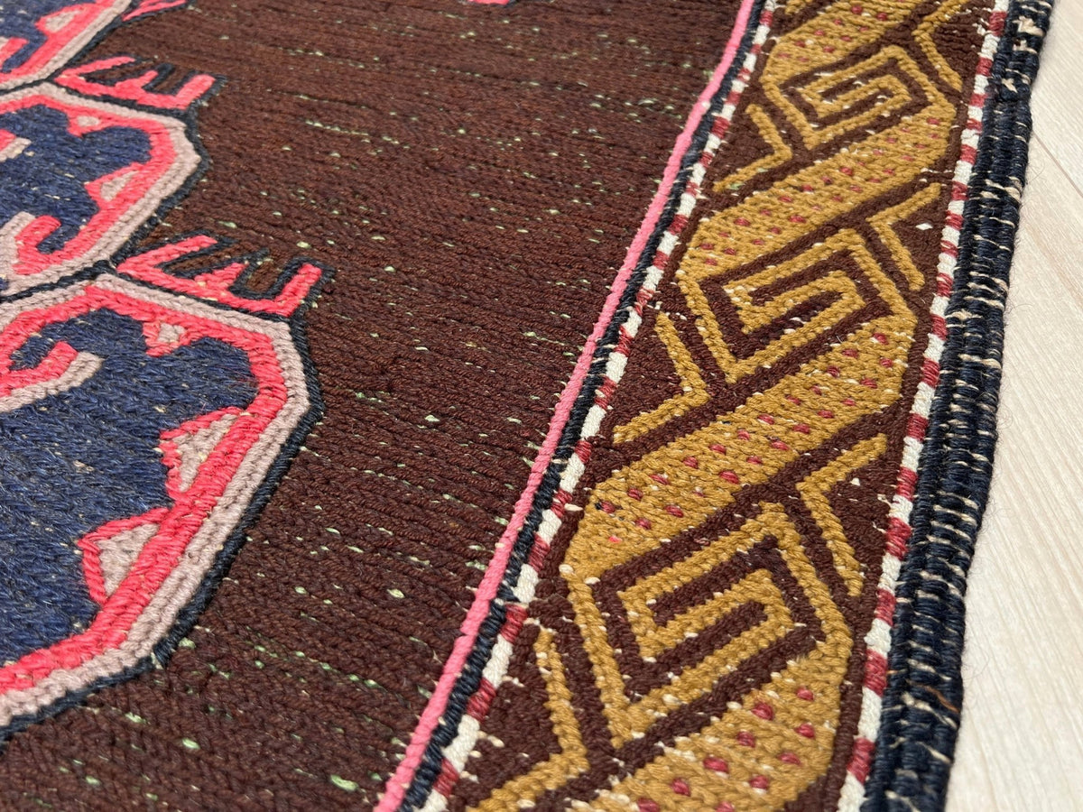 Close-up of a woven rug showing a dark brown field with a blue and pink motif on the left and a gold geometric border with pink, white, and black accents on the right.