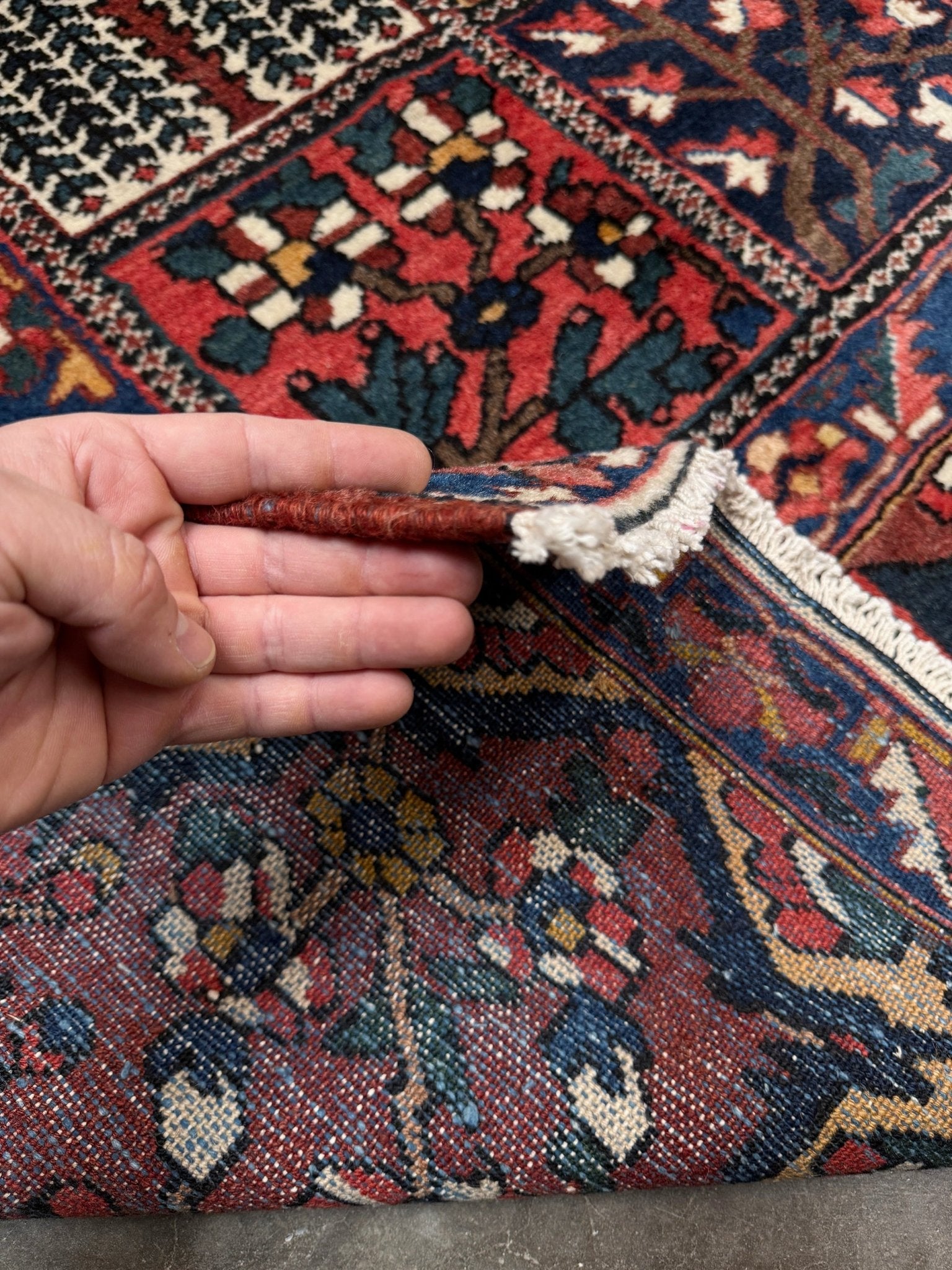 Hand holding a corner of a patterned rug with intricate designs. 10x13 handmade Persian Bakhtiari vintage rug with garden panel design, floral motifs, and red and navy tones — shown in living room at Indigo Rugs, Burlingame — San Francisco Bay Area with free shipping