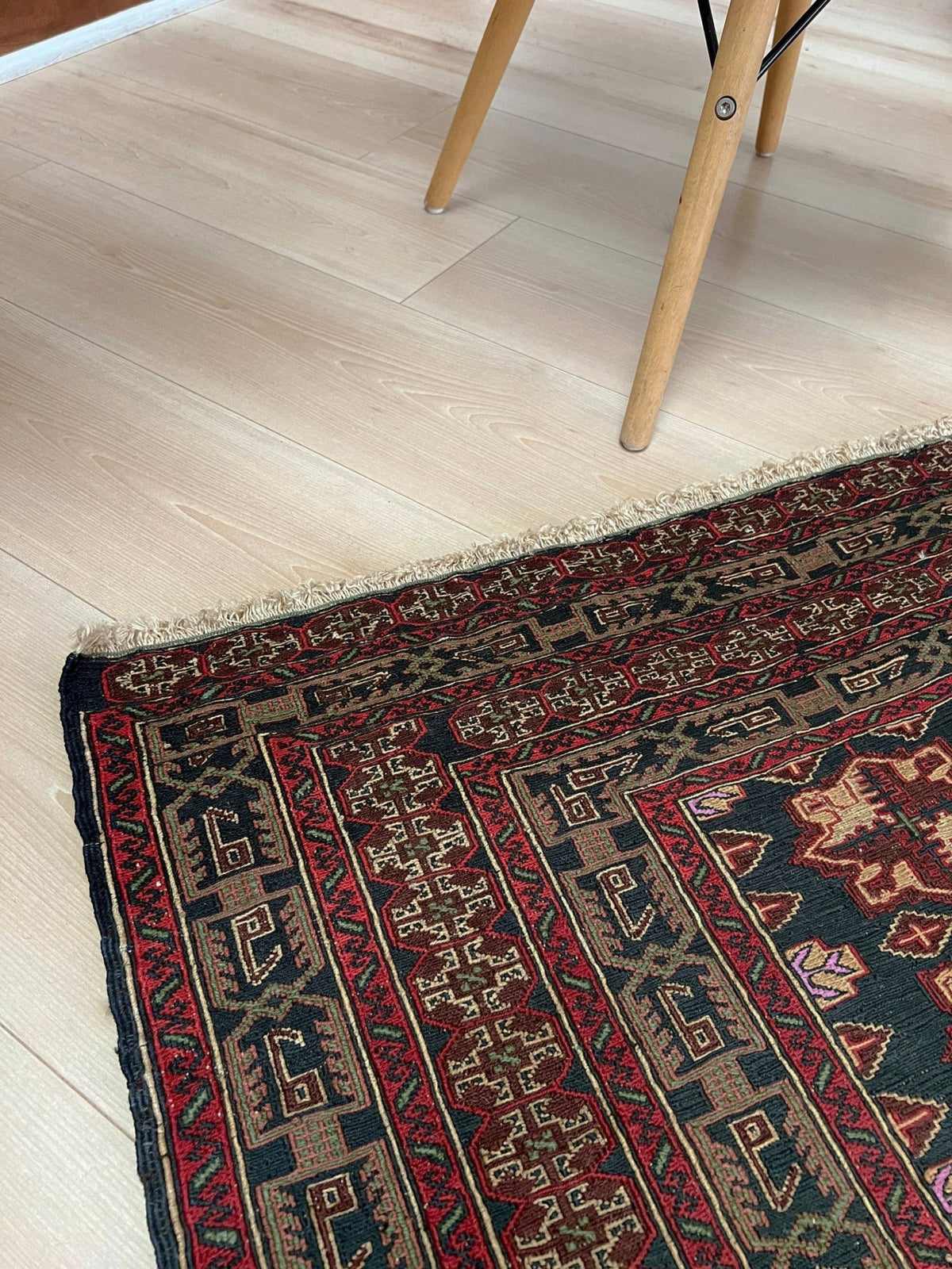 A close-up of a traditional patterned area rug with red, black, and beige geometric designs, shown on a light wood floor with the legs of a wooden chair visible in the upper frame.