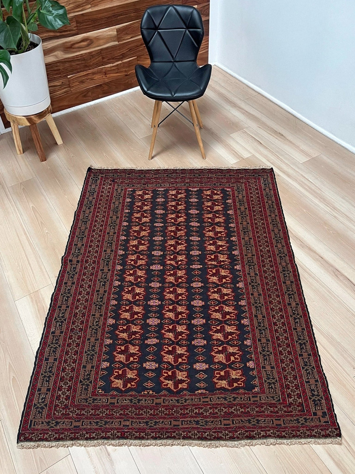 A rectangular area rug with a dark navy blue center and intricate red, brown, and beige geometric patterns, displayed on a light wood floor in a room setting with a black chair and wooden wall in the background.