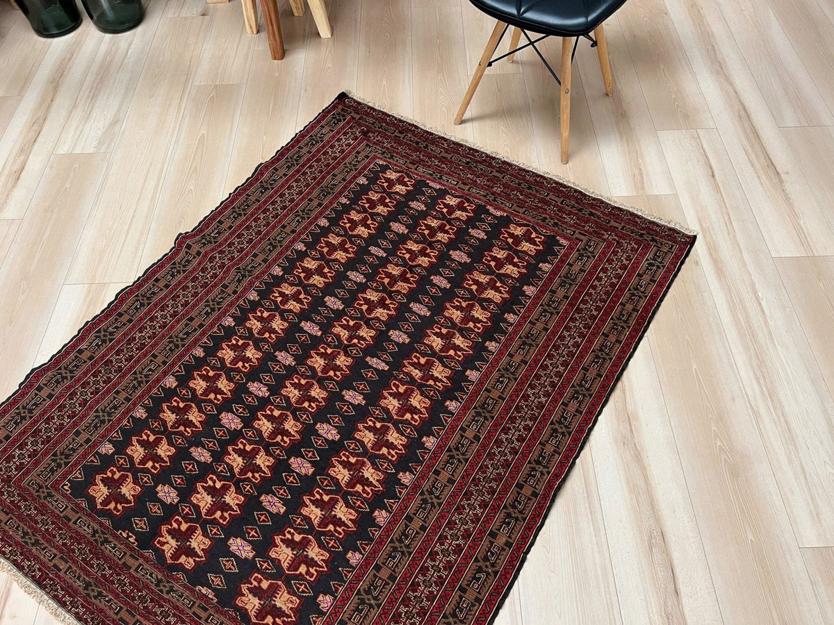 A rectangular area rug with a traditional geometric pattern in black, red, and brown, laid out on a light wood floor next to a black chair.