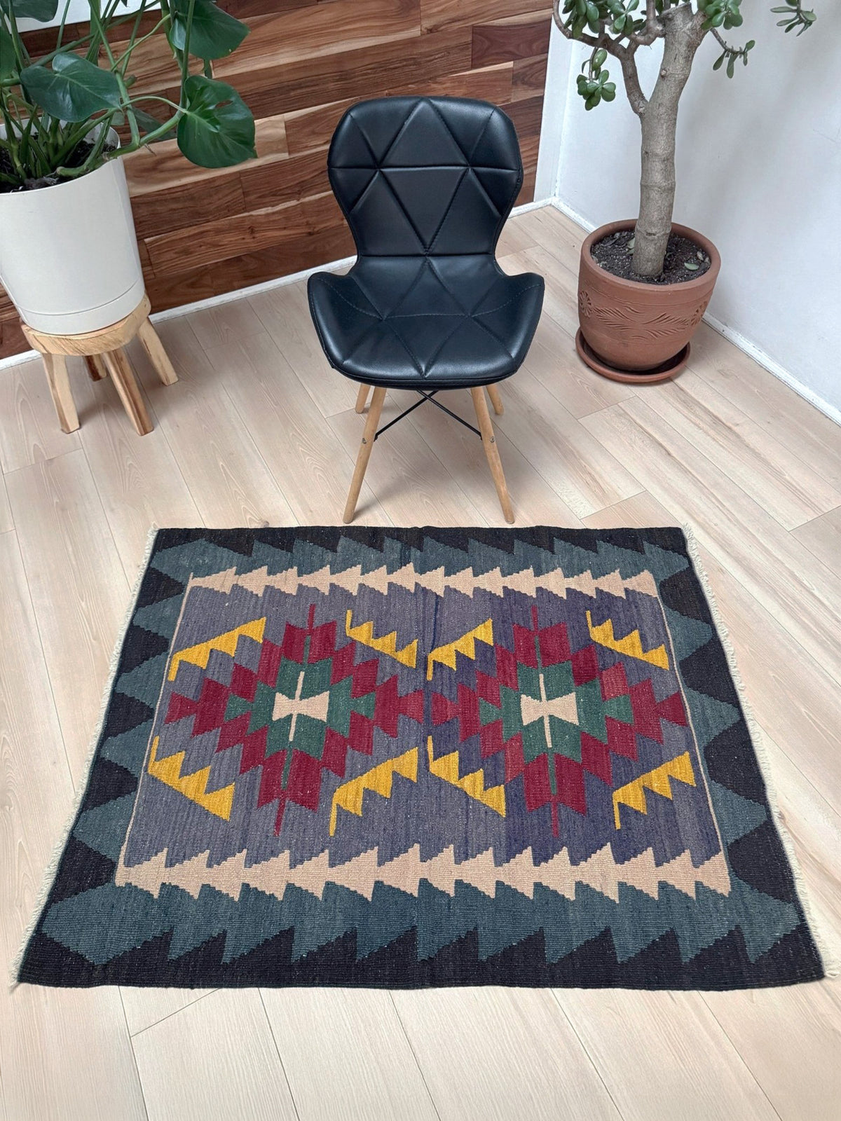 A rectangular area rug with a Southwestern geometric pattern in dark gray, red, yellow, green, and beige, styled on a light wood floor in a room with a black chair and potted plants.