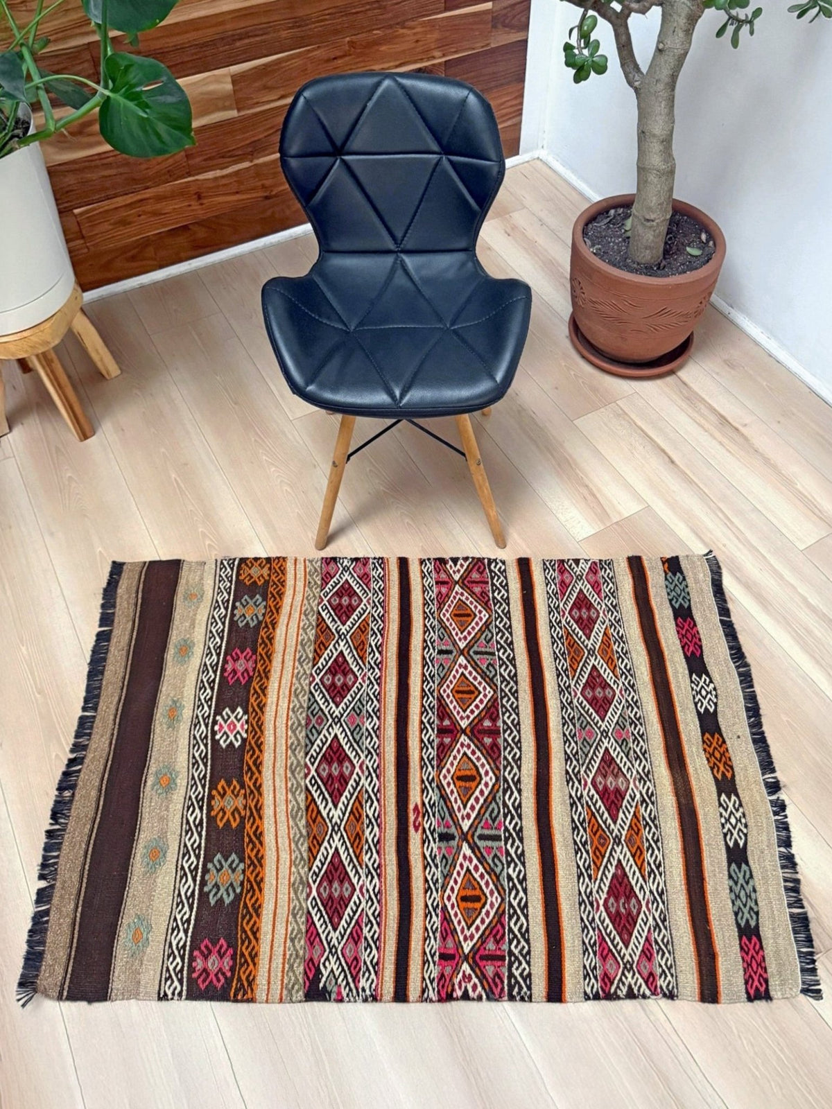 A rectangular area rug with a colorful Southwestern geometric pattern and fringe, displayed on a light wood floor in a room setting with a black chair and potted plants.