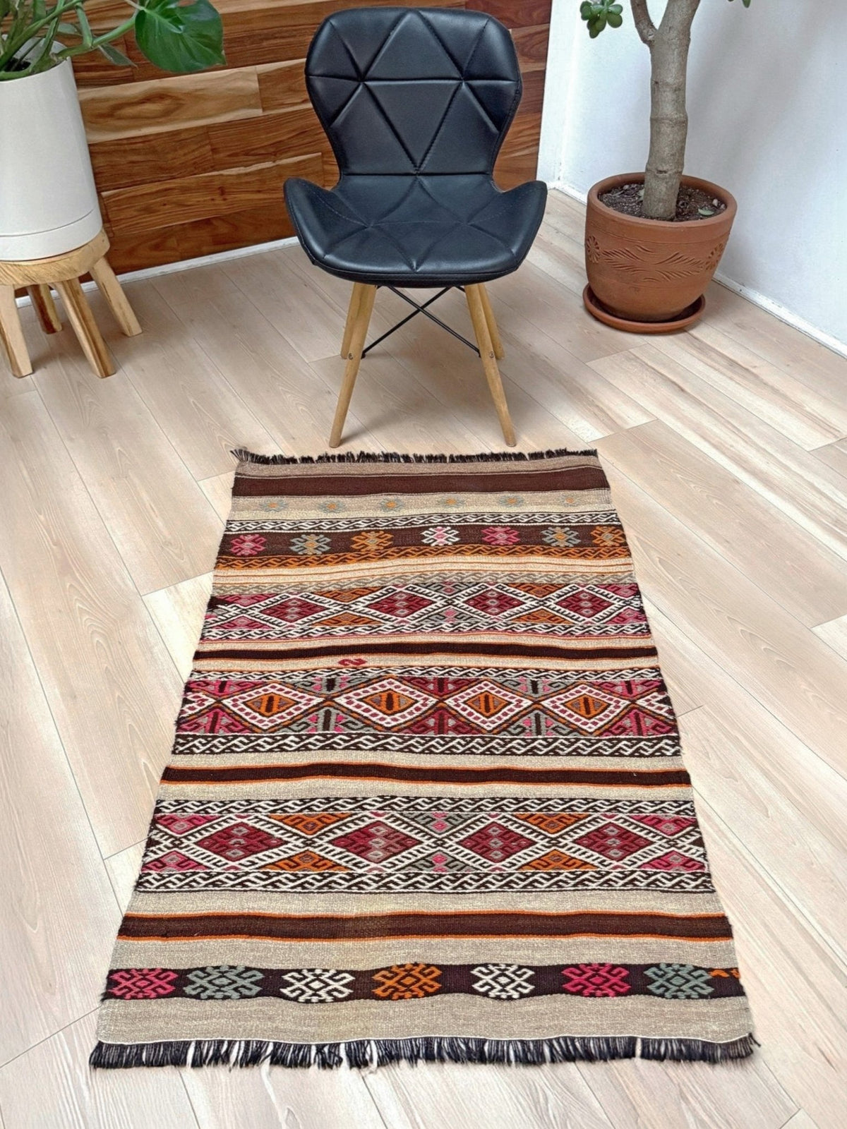 A rectangular woven area rug with a multicolor geometric kilim pattern and fringe, displayed on a light wood floor in a modern room setting with a black chair and potted plants.