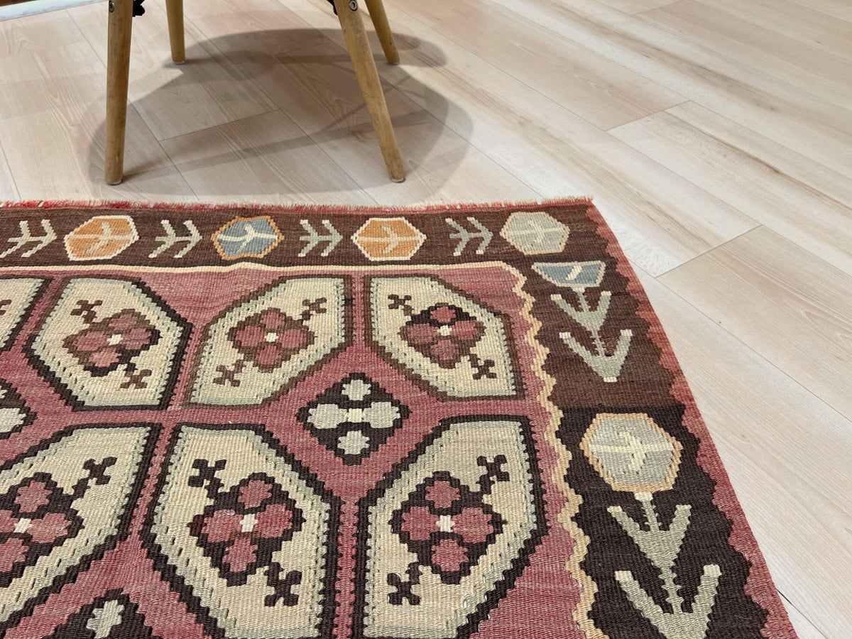 A close-up of a patterned area rug with geometric and floral motifs in dusty pink, cream, brown, and muted orange, shown on a light wood floor with the legs of a chair visible at the top.