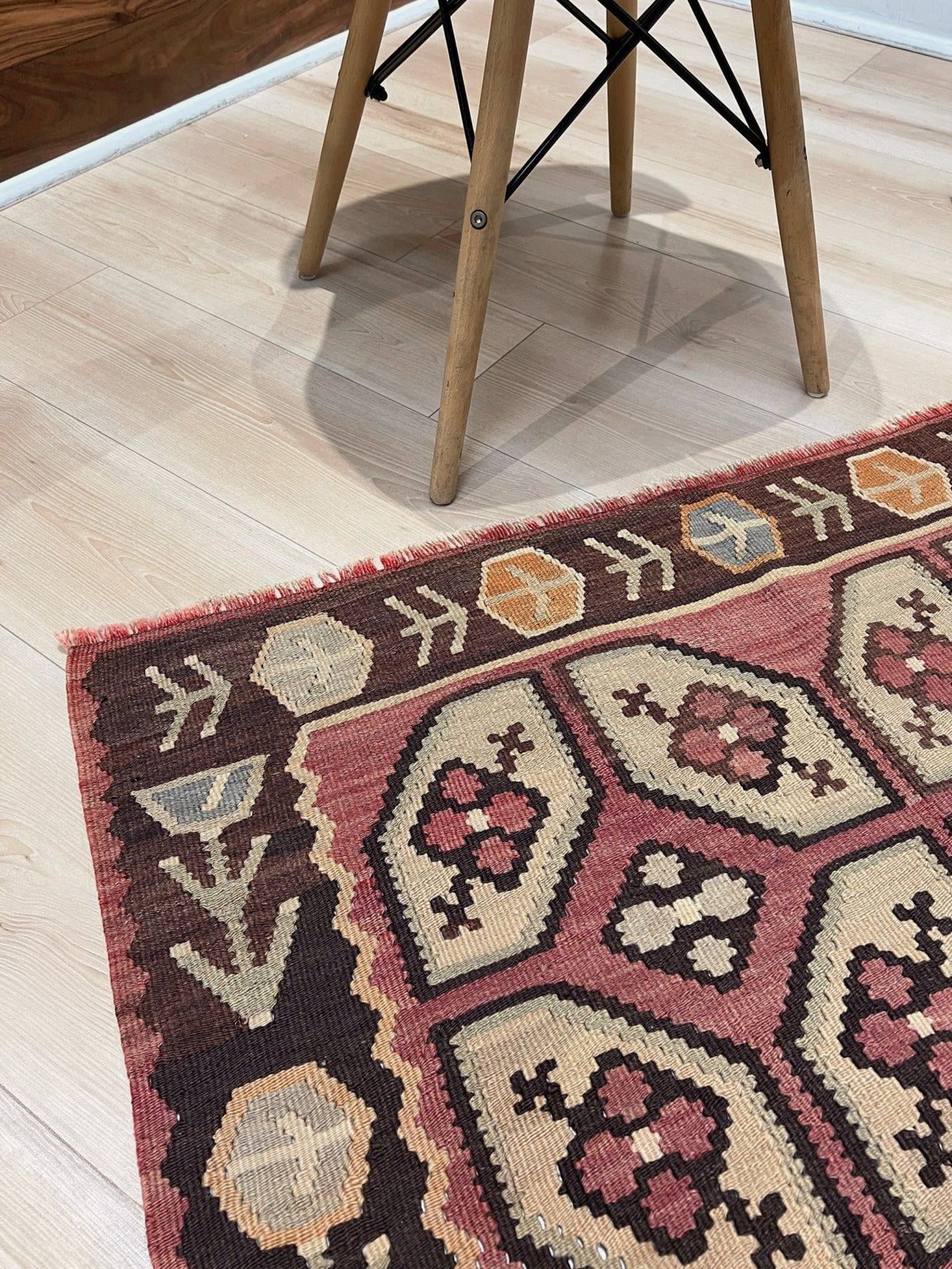 A close-up of a traditional kilim area rug with geometric patterns in dusty pink, beige, and dark brown, shown on a light wood floor next to the legs of a wooden chair.