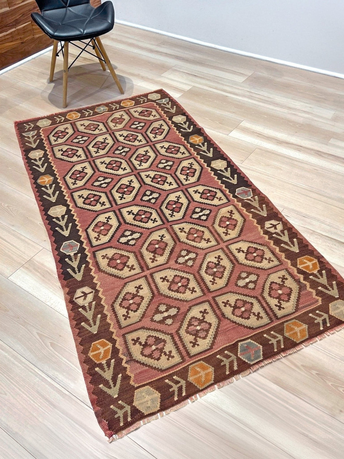 A rectangular vintage-style area rug with a geometric pattern in terracotta, beige, and dark brown, laid out on a light wood floor next to a black chair.
