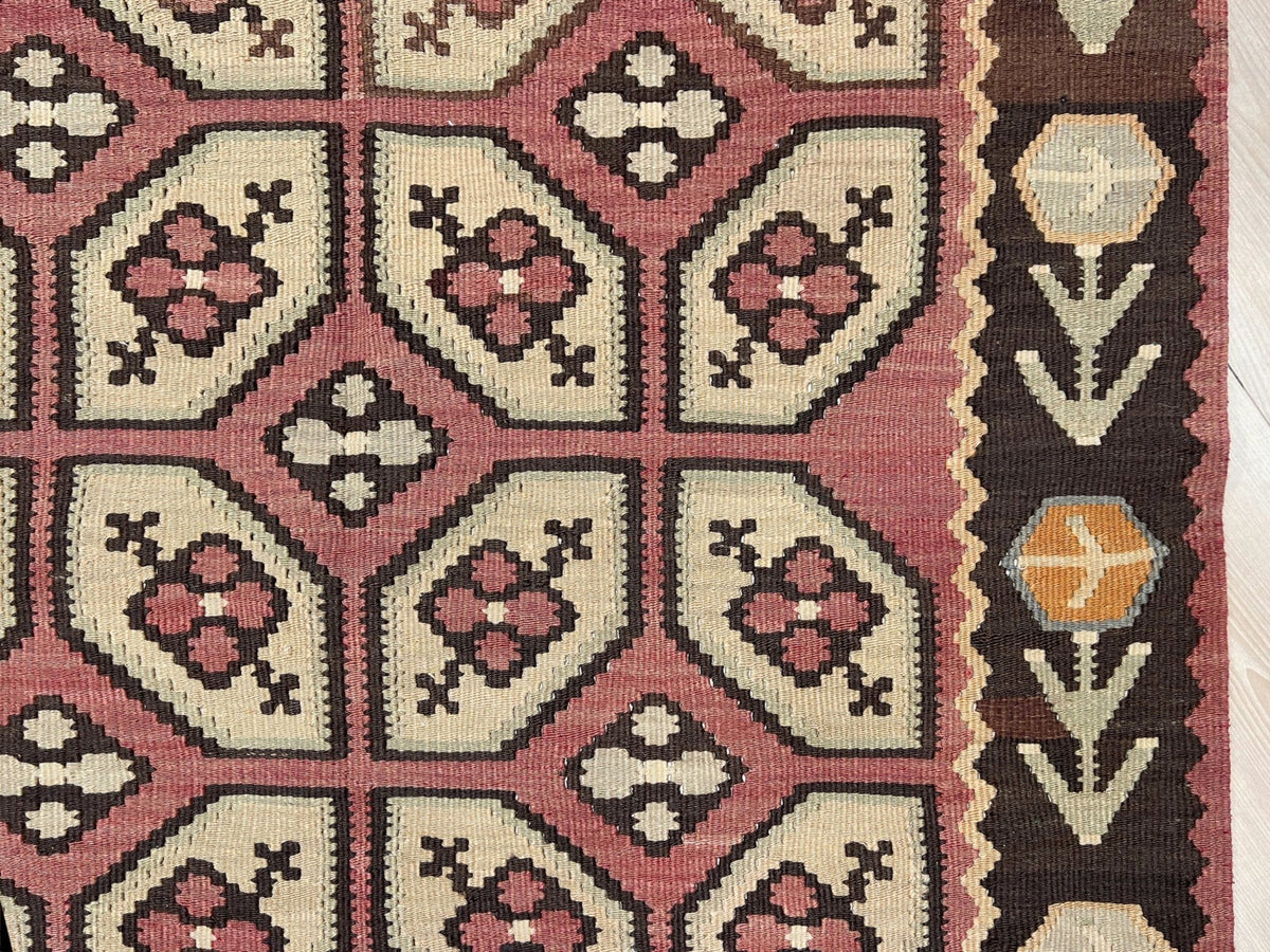 Close-up of a woven rug showing a geometric pattern with pink, cream, dark brown, and orange sections, including a decorative border on the right side.