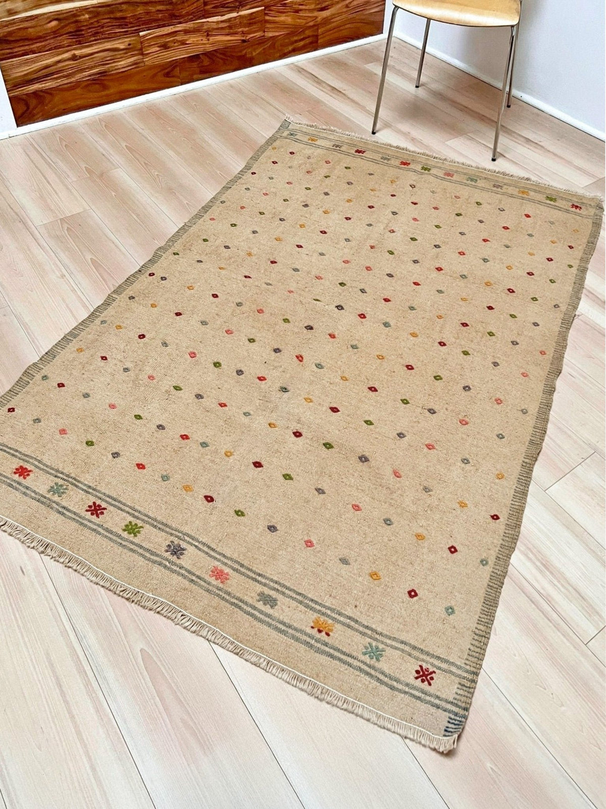 A rectangular beige area rug with a scattered multicolor dot pattern and a decorative border, shown on a light wood plank floor in a room setting.