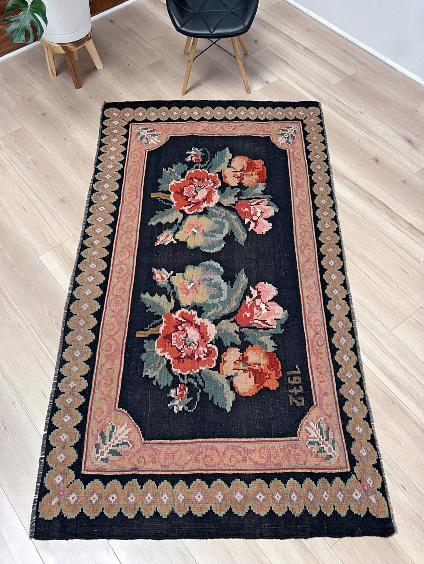 Decorative floral rug on a wooden floor with a chair in the background. vintage moldovan rose kilim rug 4x7 handmade floral black field dated 1972 handmade rug san francisco bay area rug shop indigo rugs