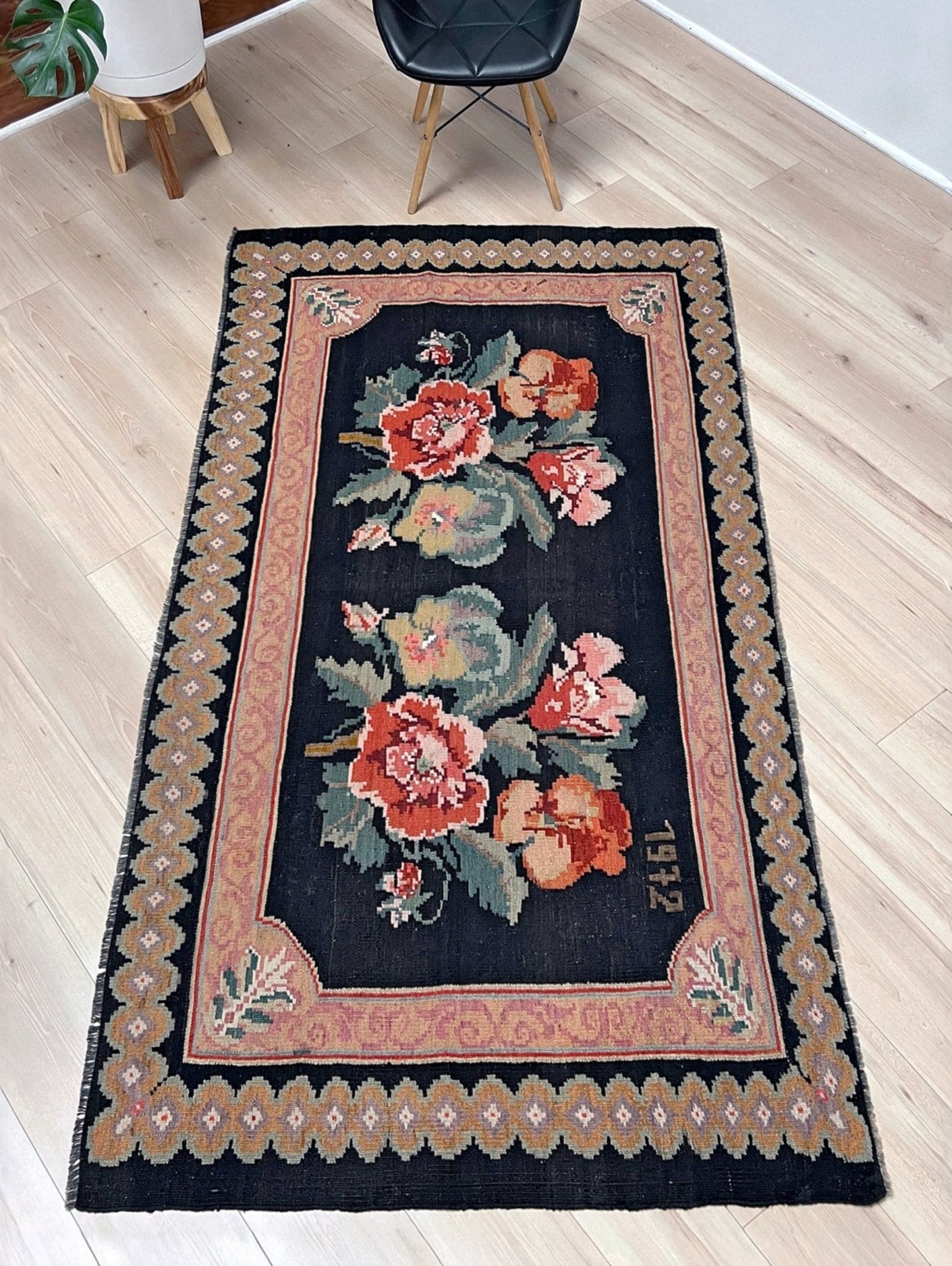 Decorative floral rug on a wooden floor with a chair in the background. vintage moldovan rose kilim rug 4x7 handmade floral black field dated 1972 handmade rug san francisco bay area rug shop indigo rugs