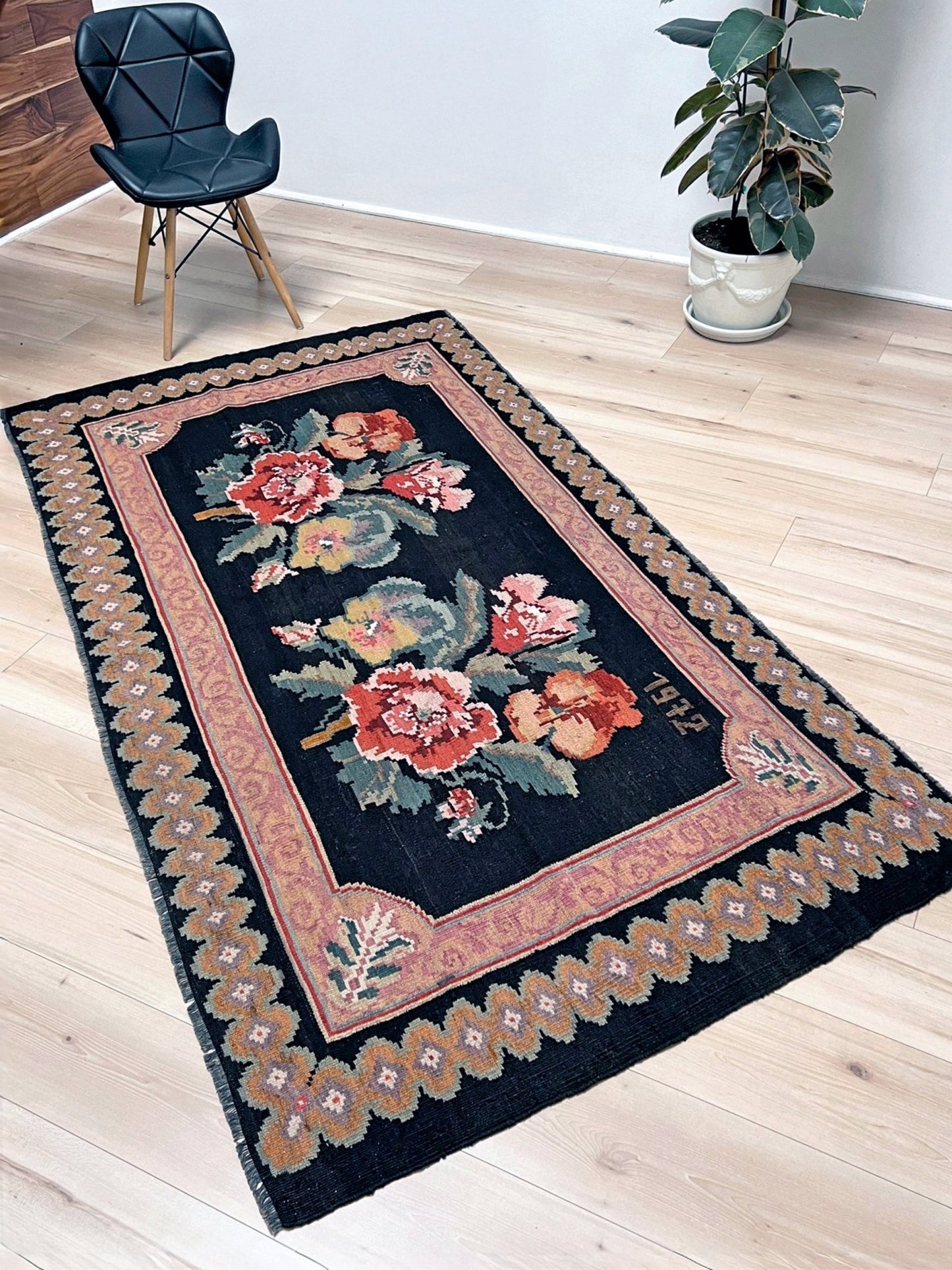 Decorative floral rug on a wooden floor with a chair and plant in the background. vintage moldovan rose kilim rug 4x7 handmade floral black field dated 1972 handmade rug san francisco bay area rug shop indigo rugs