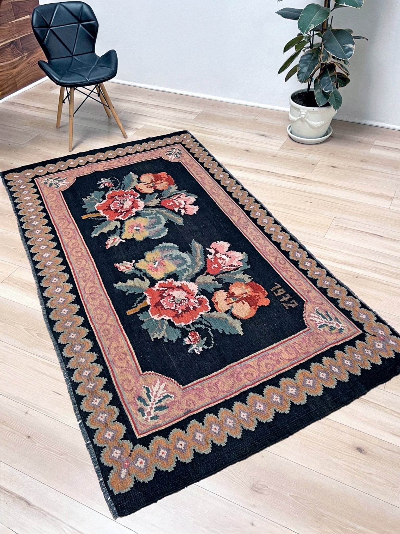 Decorative floral rug on a wooden floor with a chair and plant in the background. vintage moldovan rose kilim rug 4x7 handmade floral black field dated 1972 handmade rug san francisco bay area rug shop indigo rugs