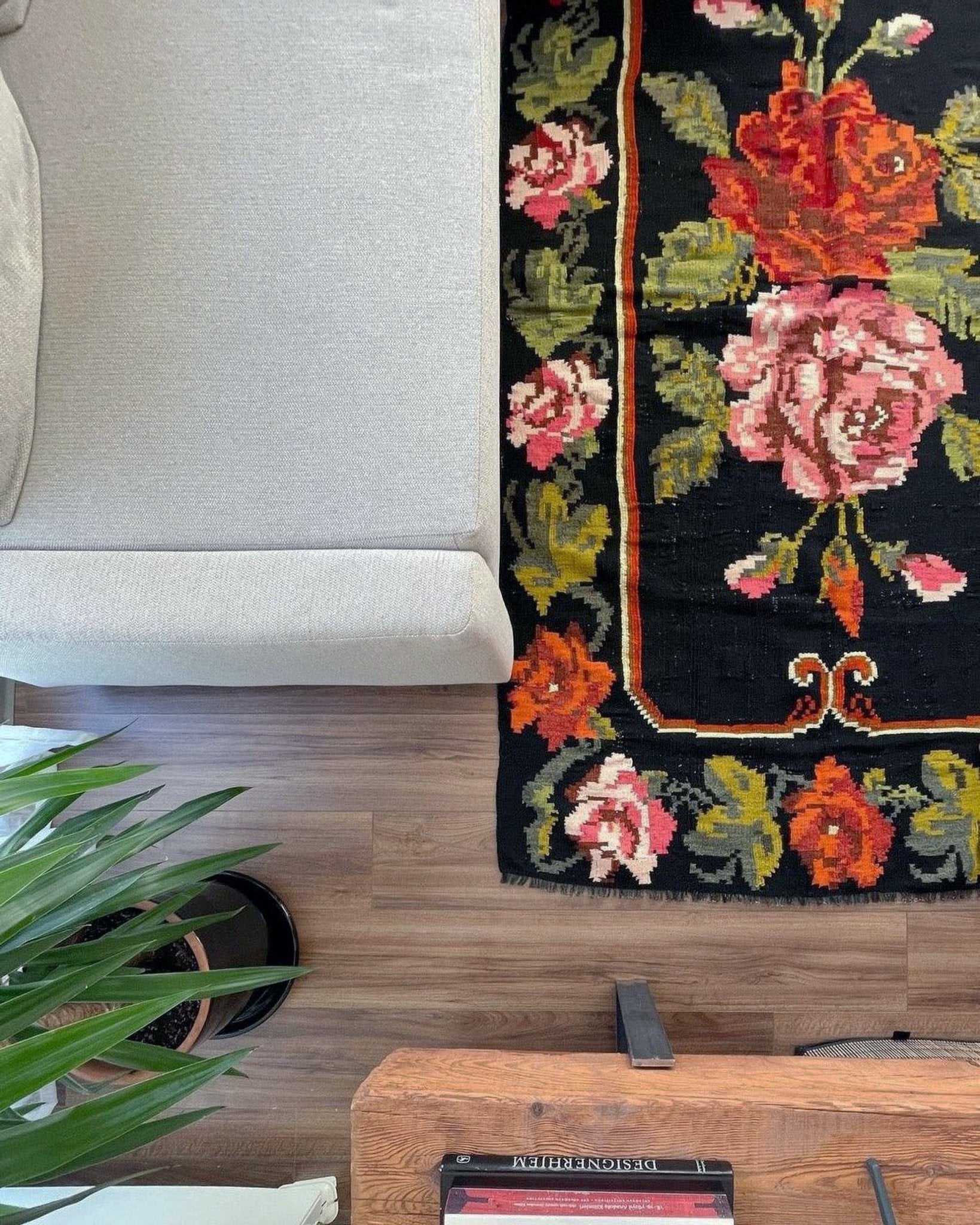 Floral patterned rug on a wooden floor next to a beige sofa and a plant. Vintage moldovan floral kilim rug 5x8 handmade rose design black background flatweave 1960s eastern european wool rug san francisco bay area rug shop indigo rugs