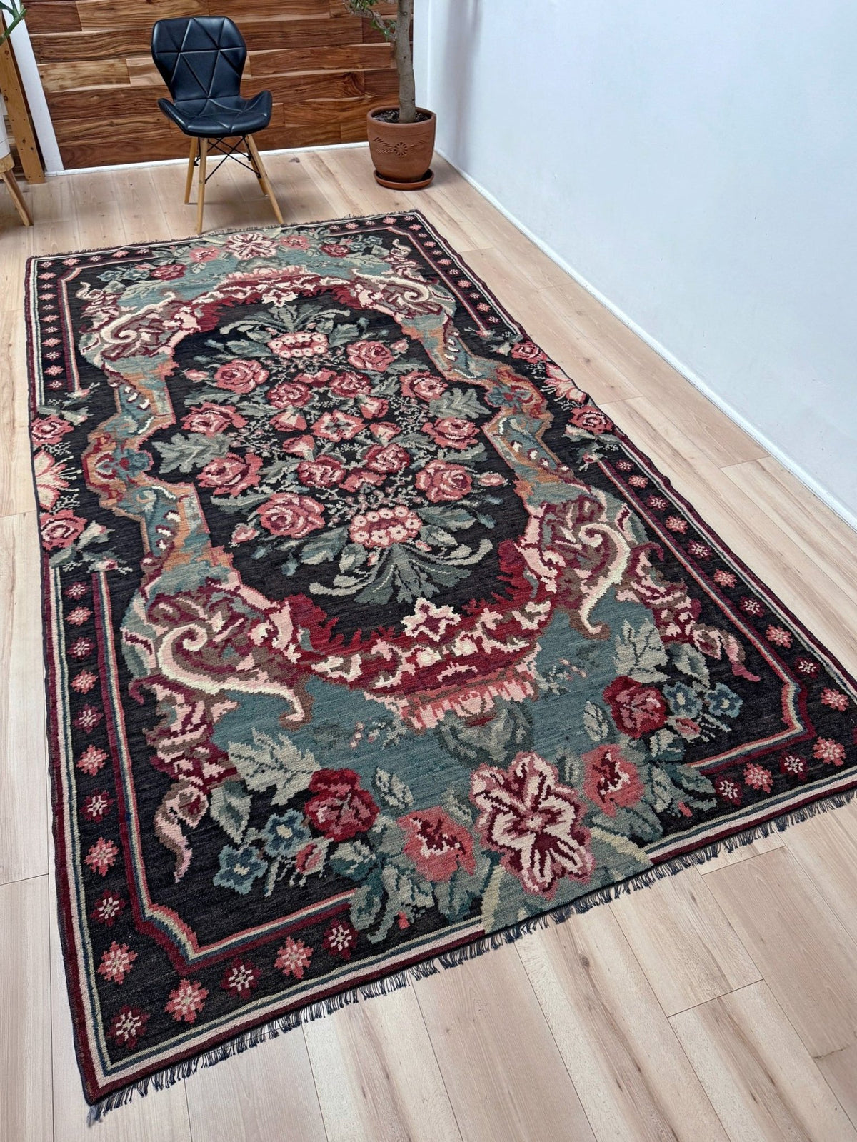 A rectangular vintage-style area rug with an ornate floral medallion pattern, displayed on a light wood floor in a room setting. Vintage Moldovan rose kilim floral rug handmade wool flatweave 6x10  from Indigo Rugs Burlingame Bay Area.
