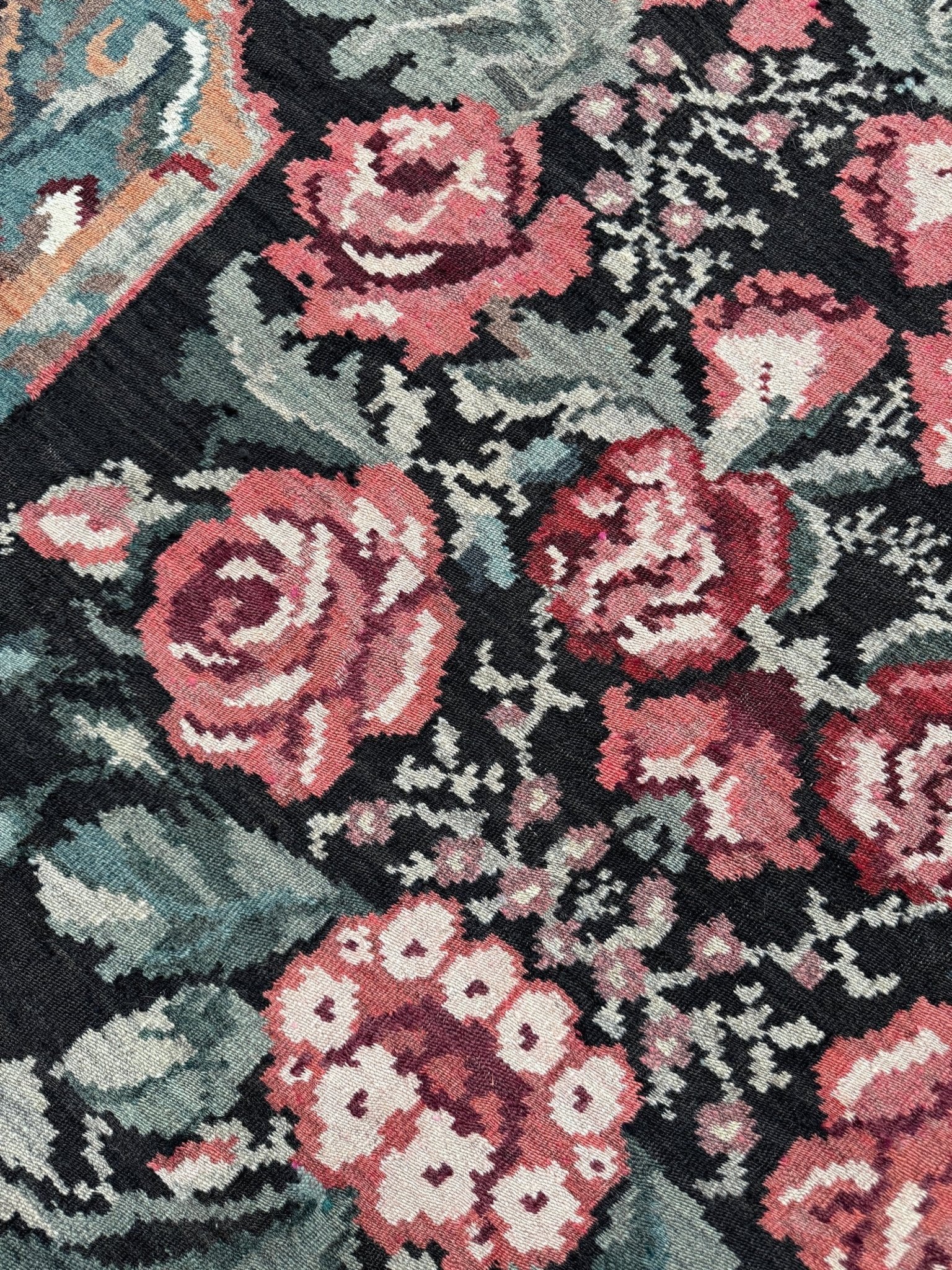 Close-up of a floral patterned textile with pink and coral roses, muted green leaves, and a dark charcoal background. Vintage Moldovan rose kilim floral rug handmade wool flatweave 6x10  from Indigo Rugs Burlingame Bay Area.