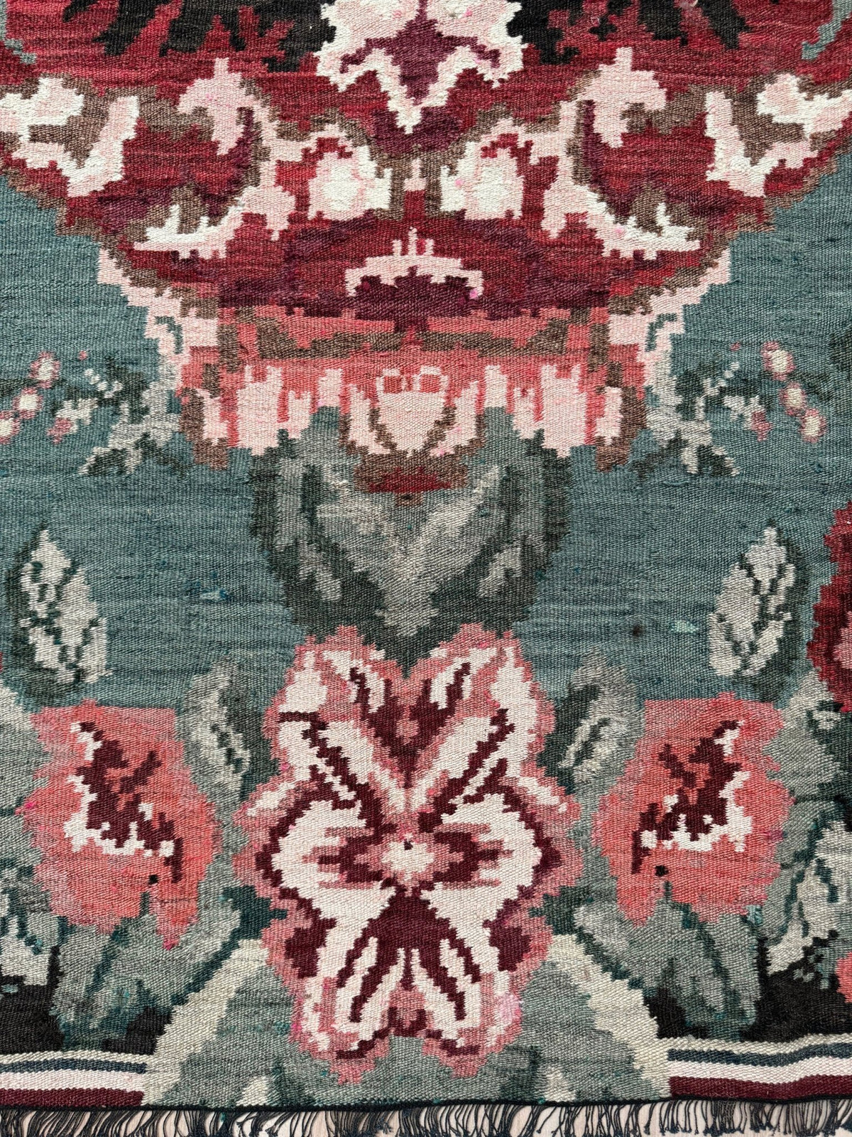Close-up of a patterned rug with a muted teal-blue background and ornate floral motifs in burgundy, dusty pink, cream, and gray, with fringe visible along the bottom edge. Vintage Moldovan rose kilim floral rug handmade wool flatweave 6x10  from Indigo Rugs Burlingame Bay Area.