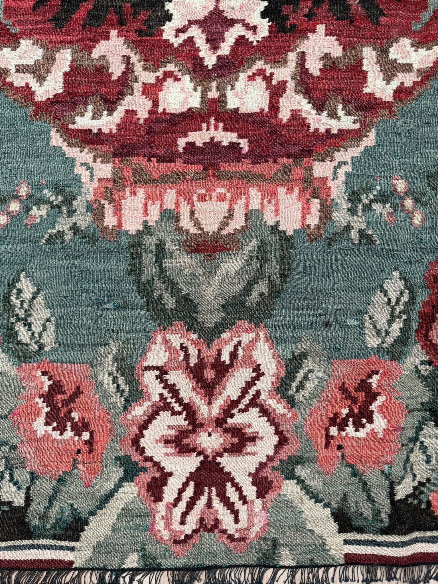 Close-up of a patterned rug with a muted teal-blue background and ornate floral motifs in burgundy, dusty pink, cream, and gray, with fringe visible along the bottom edge. Vintage Moldovan rose kilim floral rug handmade wool flatweave 6x10  from Indigo Rugs Burlingame Bay Area.